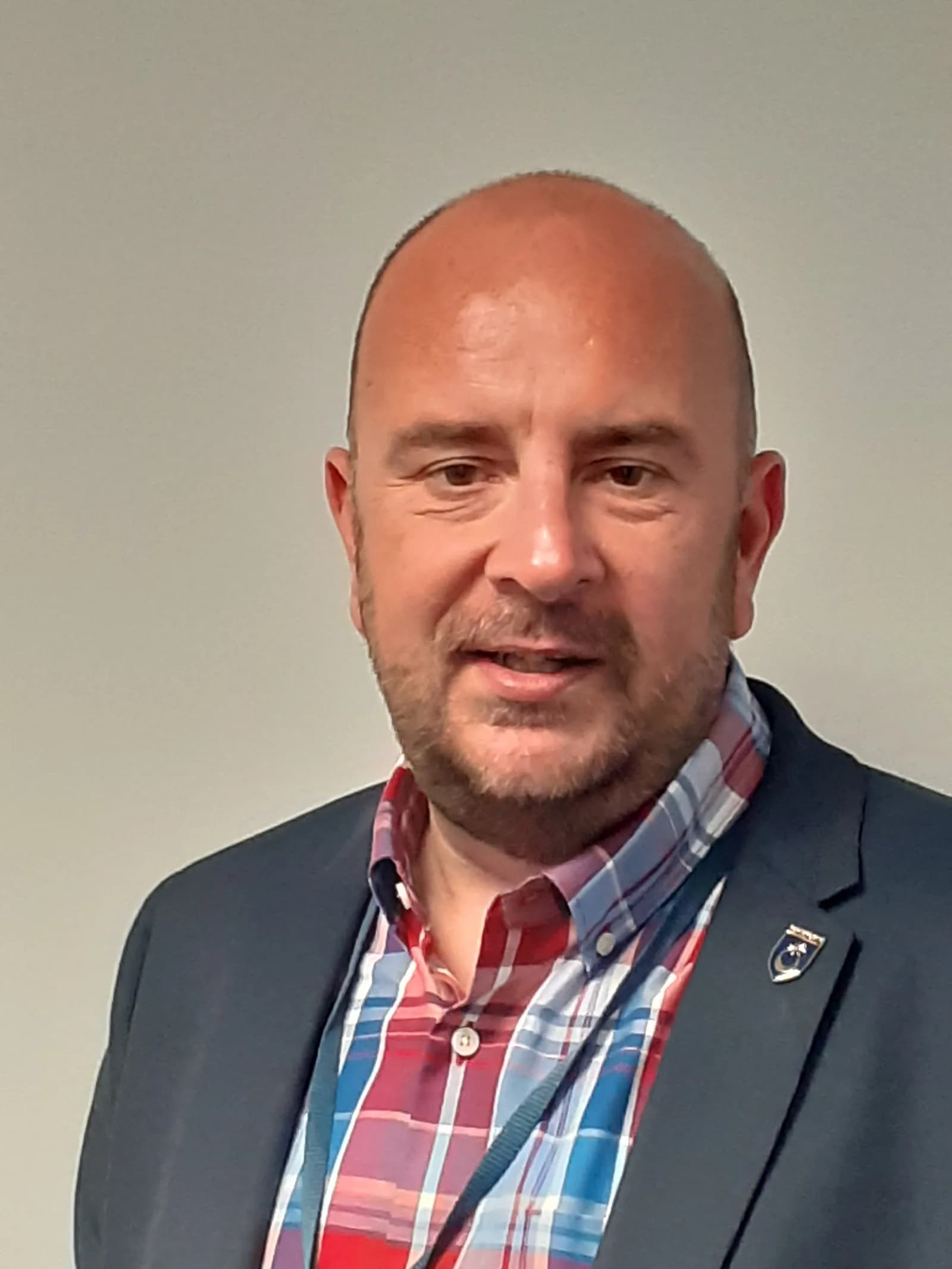 A man with a bald head and beard smiles slightly, wearing a dark blue blazer over a red, white, and blue plaid shirt with a blue lanyard.
