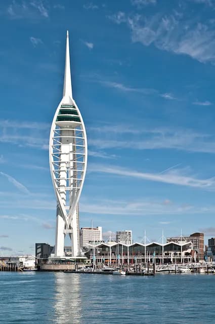 A modern white tower with a pointed top stands tall against a blue sky, overlooking a body of water with buildings and boats in the background.