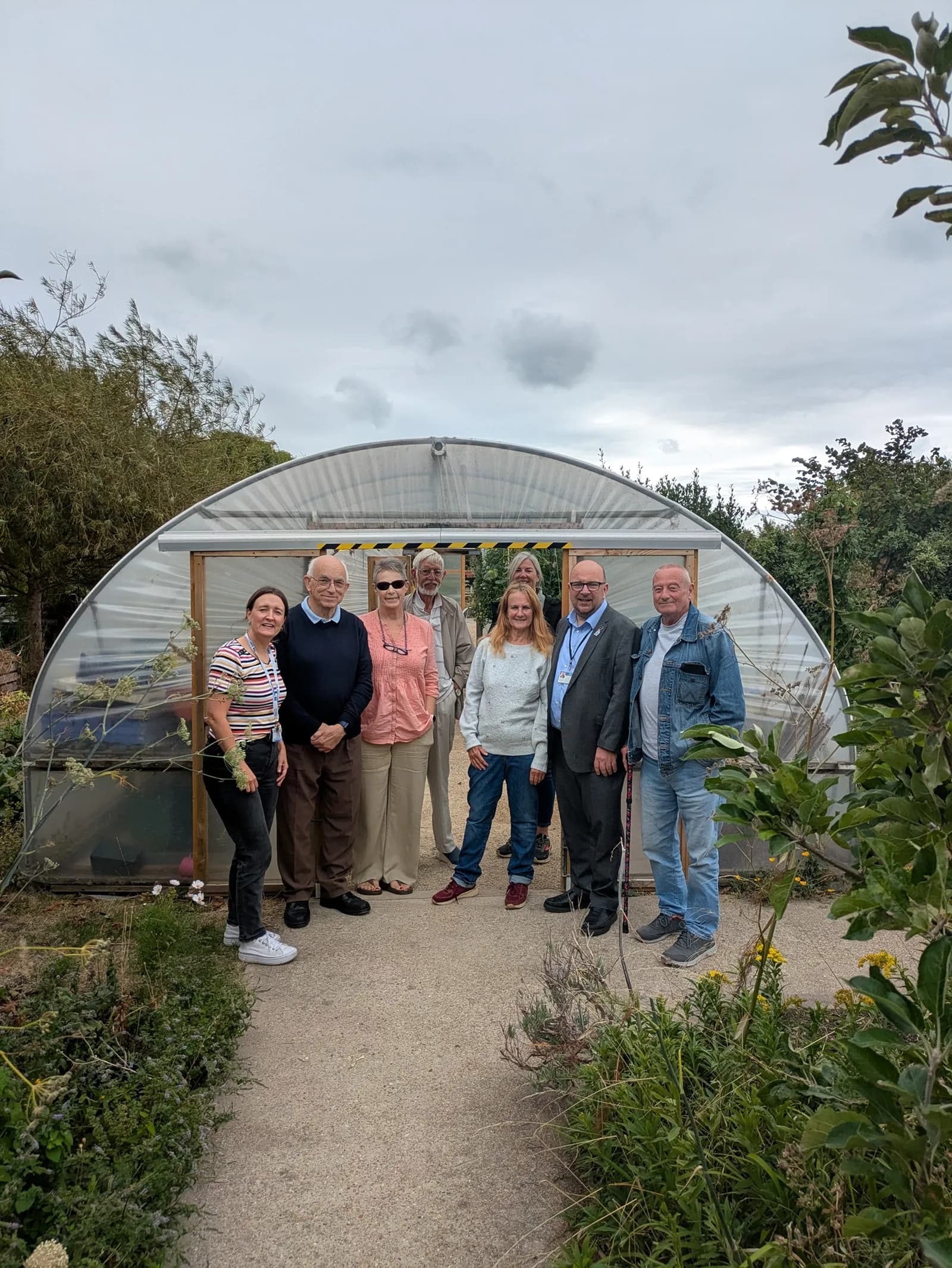 A group of seven adults and one child stand in front of a small, curved greenhouse.